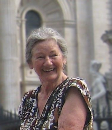 An elderly woman is smiling in front of a building.