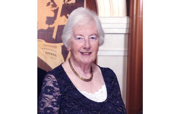 An elderly woman in a blue dress and necklace is smiling for the camera.