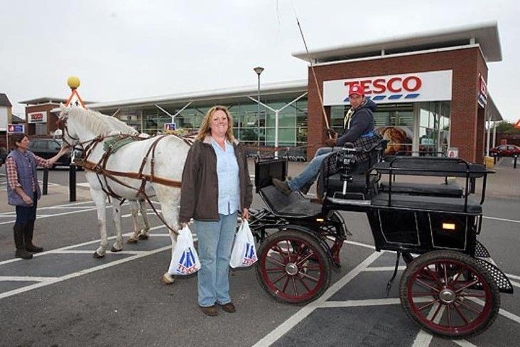 A woman stands next to a horse drawn carriage in front of a tesco store