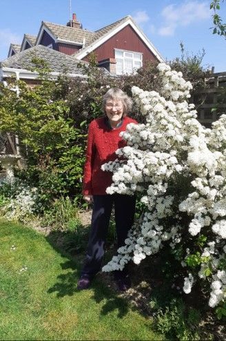 An elderly woman is standing next to a bush with white flowers in a garden.