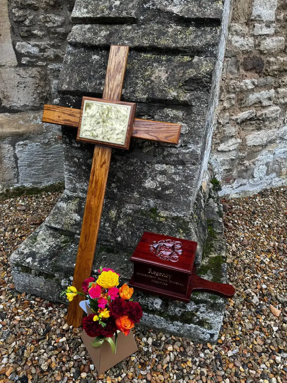 Wooden cross and box with flowers against a stone wall.