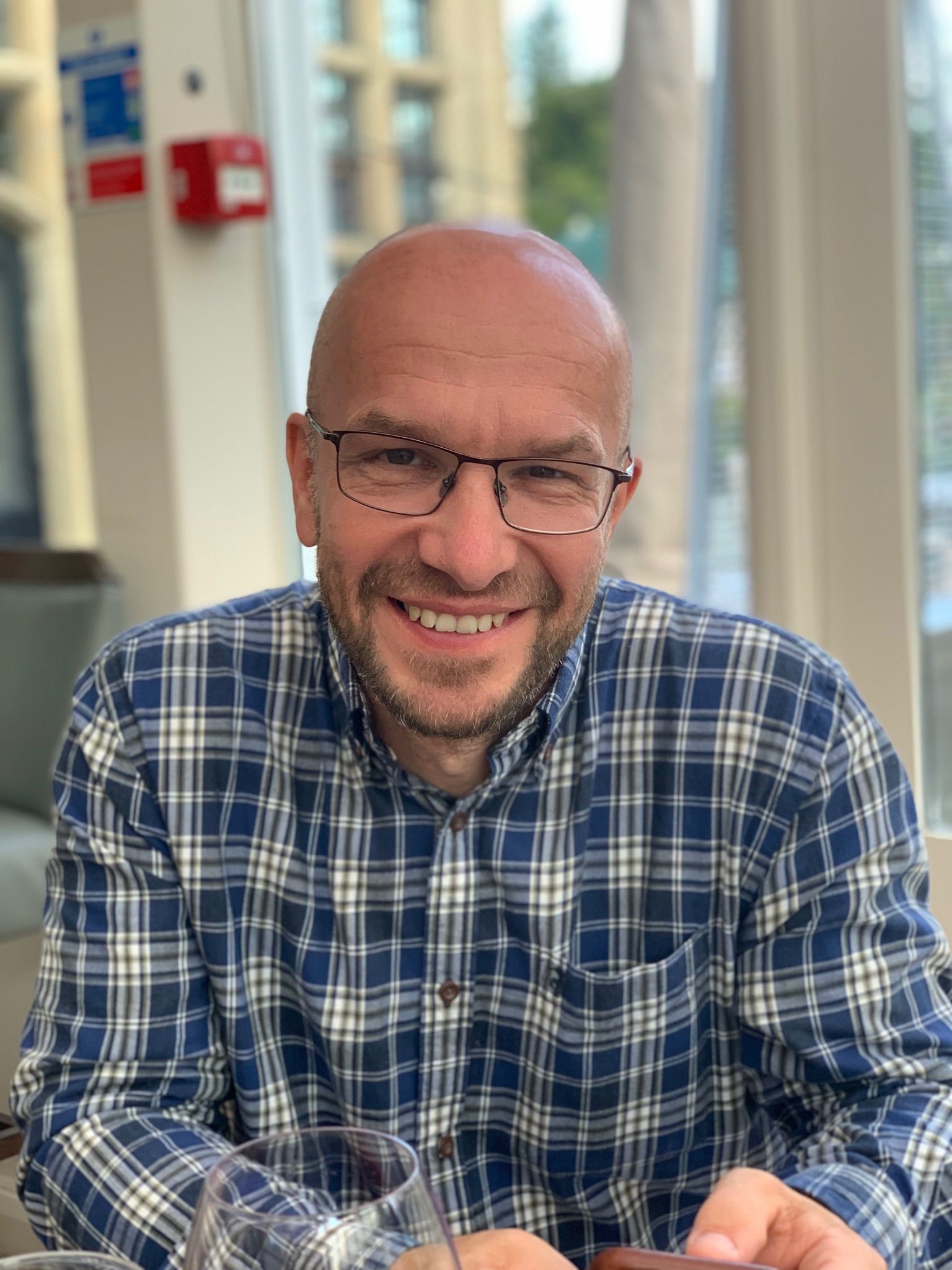 Bald man in plaid shirt and glasses smiles indoors.