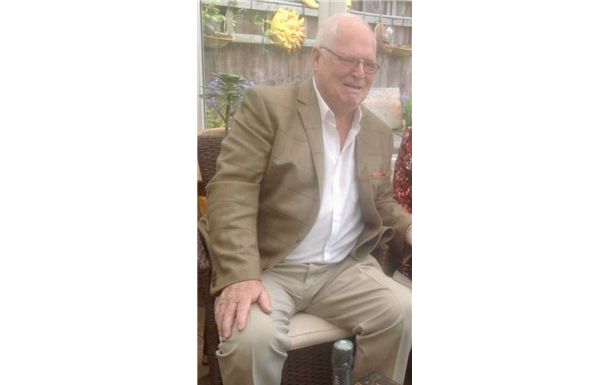 An individual wearing a tan blazer and white shirt sits outdoors, smiling softly in a garden setting.