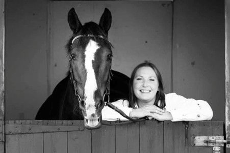 An individual wearing a tan blazer and white shirt sits outdoors, smiling softly in a garden setting.