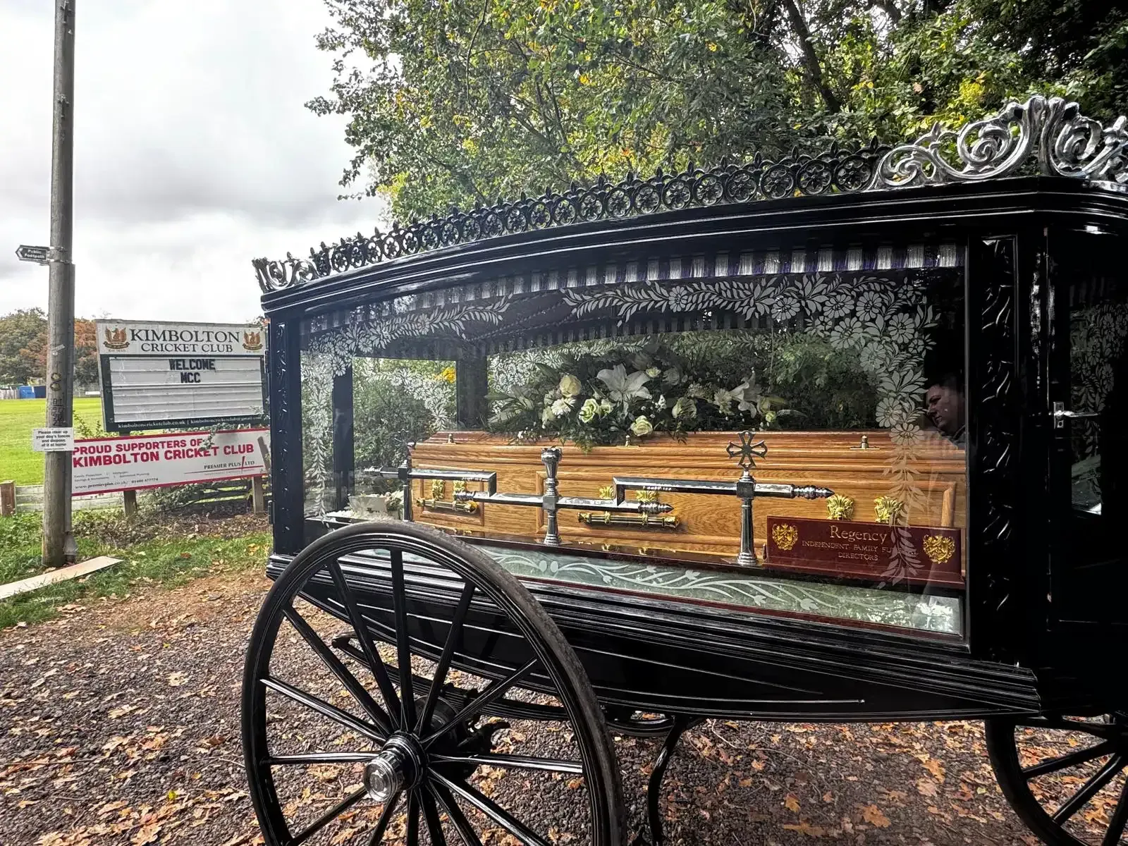 Black horse-drawn hearse with golden coffin, glass sides, and floral arrangements, outdoors.