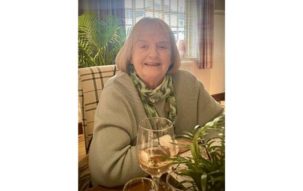 Woman with short blonde hair, wearing a green sweater and scarf, smiling, sitting at a table with wine glasses.