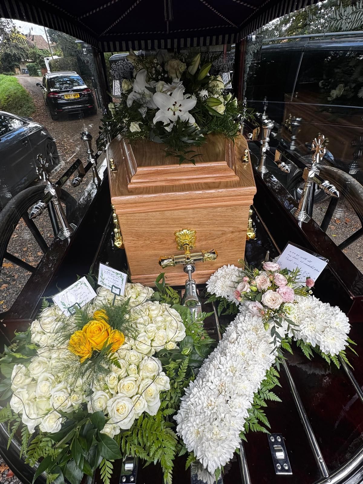 Wooden casket in a hearse, adorned with floral arrangements.