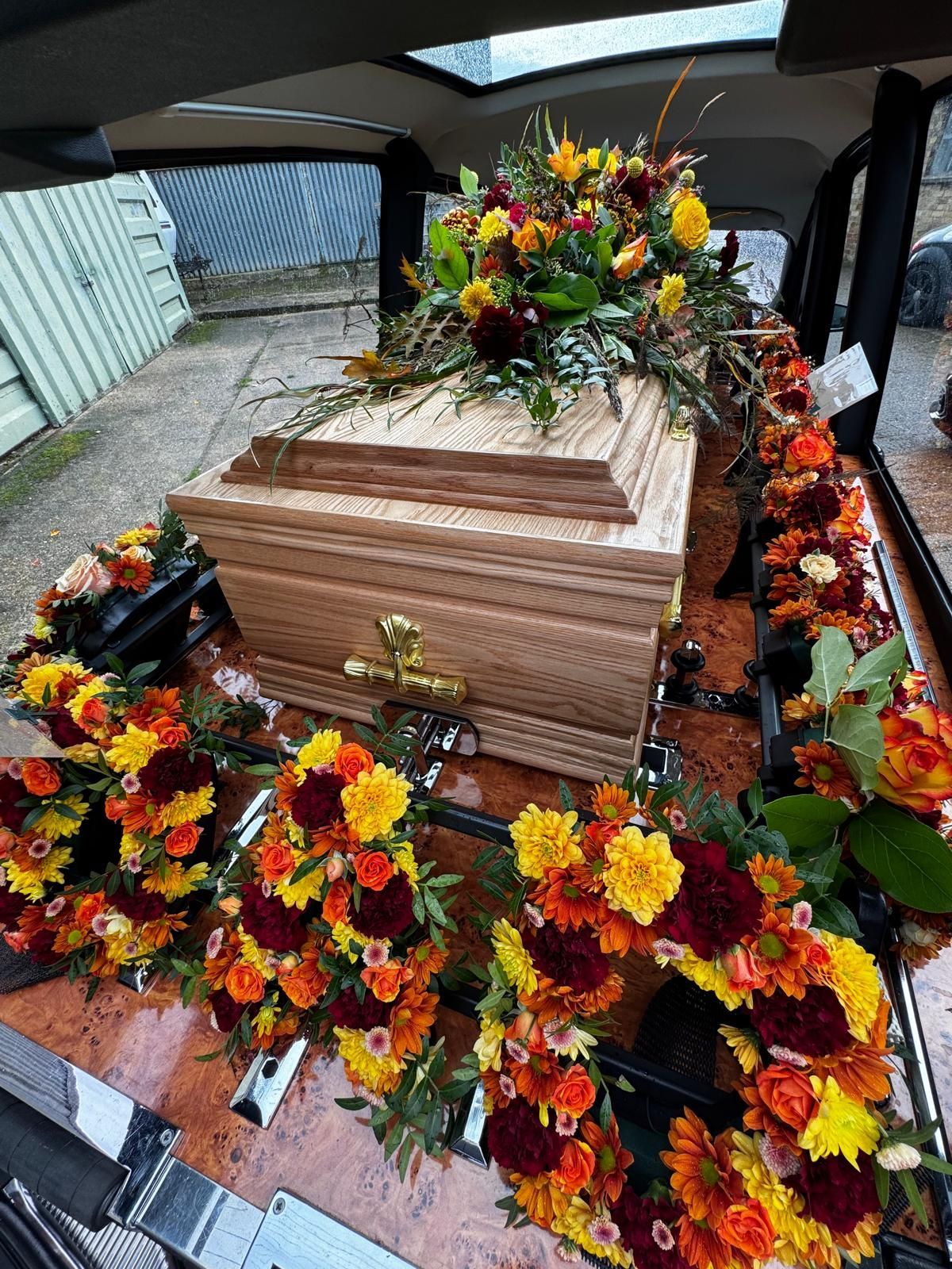 Wooden casket in a hearse, adorned with autumn-colored floral wreaths and arrangement.