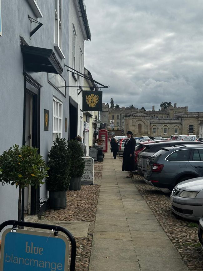 A street with a cafe, cars parked on the right, and a castle in the distance under an overcast sky.