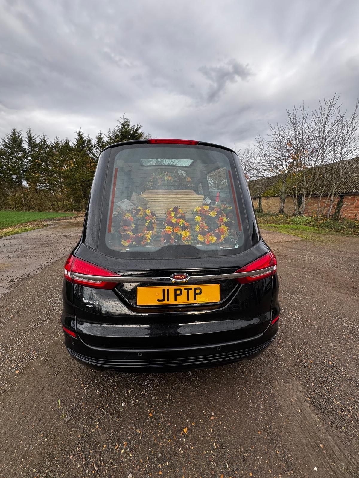 Black hearse with flowers inside, parked outside. Cloudy day.