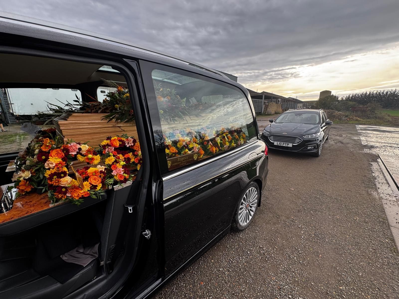 Black hearse with open door, casket with flower arrangement, parked on gravel road. Another car behind. Cloudy sky.