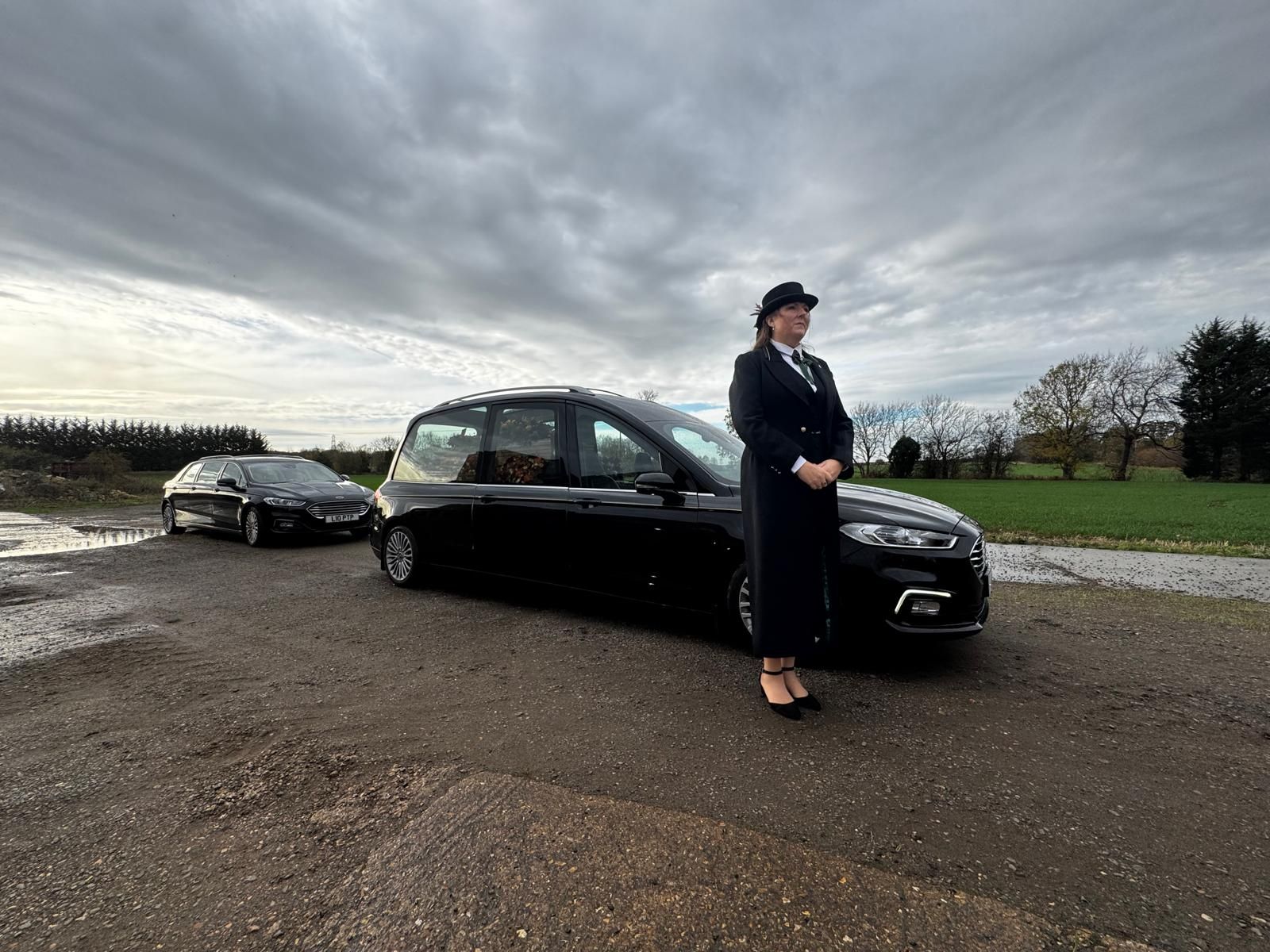 Woman in black suit stands by black car on gravel road under cloudy sky, another car in background.