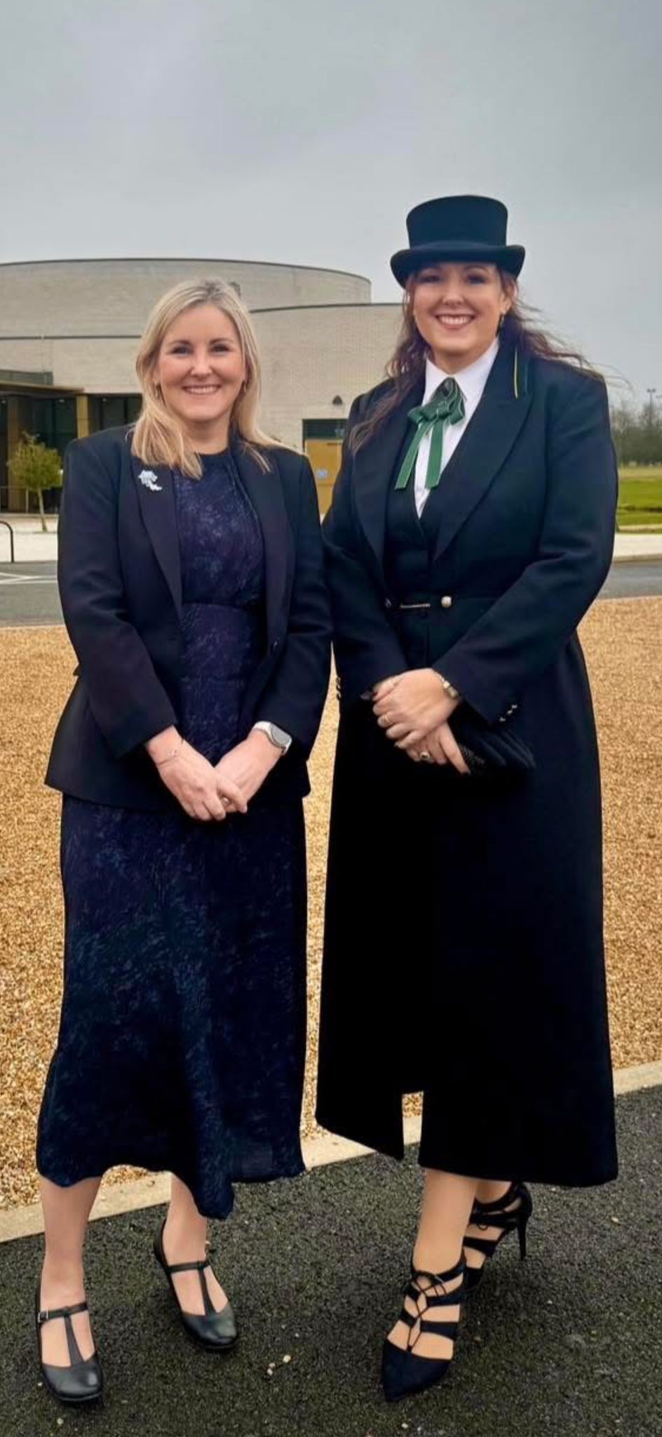 Two women, one in a top hat and coat, stand side by side outside on an overcast day.