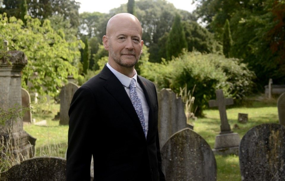 Man in a suit stands in a cemetery with tombstones and trees.