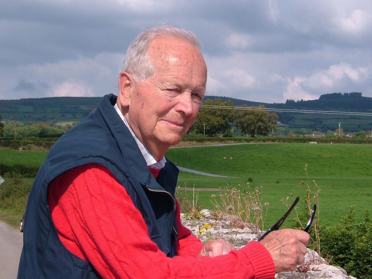 Man smiling in a red shirt and navy vest, sitting outdoors with green fields and cloudy hills behind him