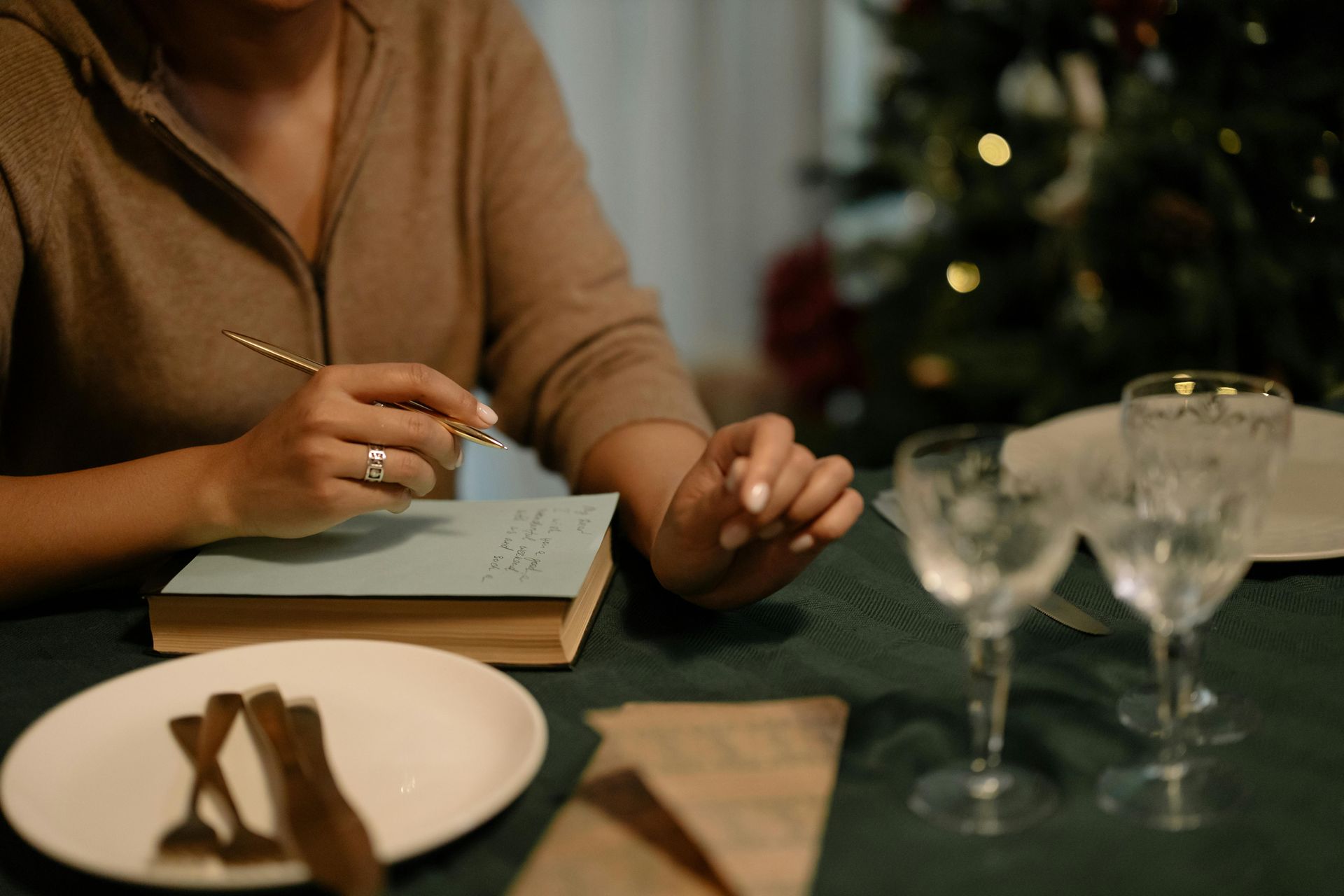 Person writing at a table set for a meal, with a Christmas tree in the background.