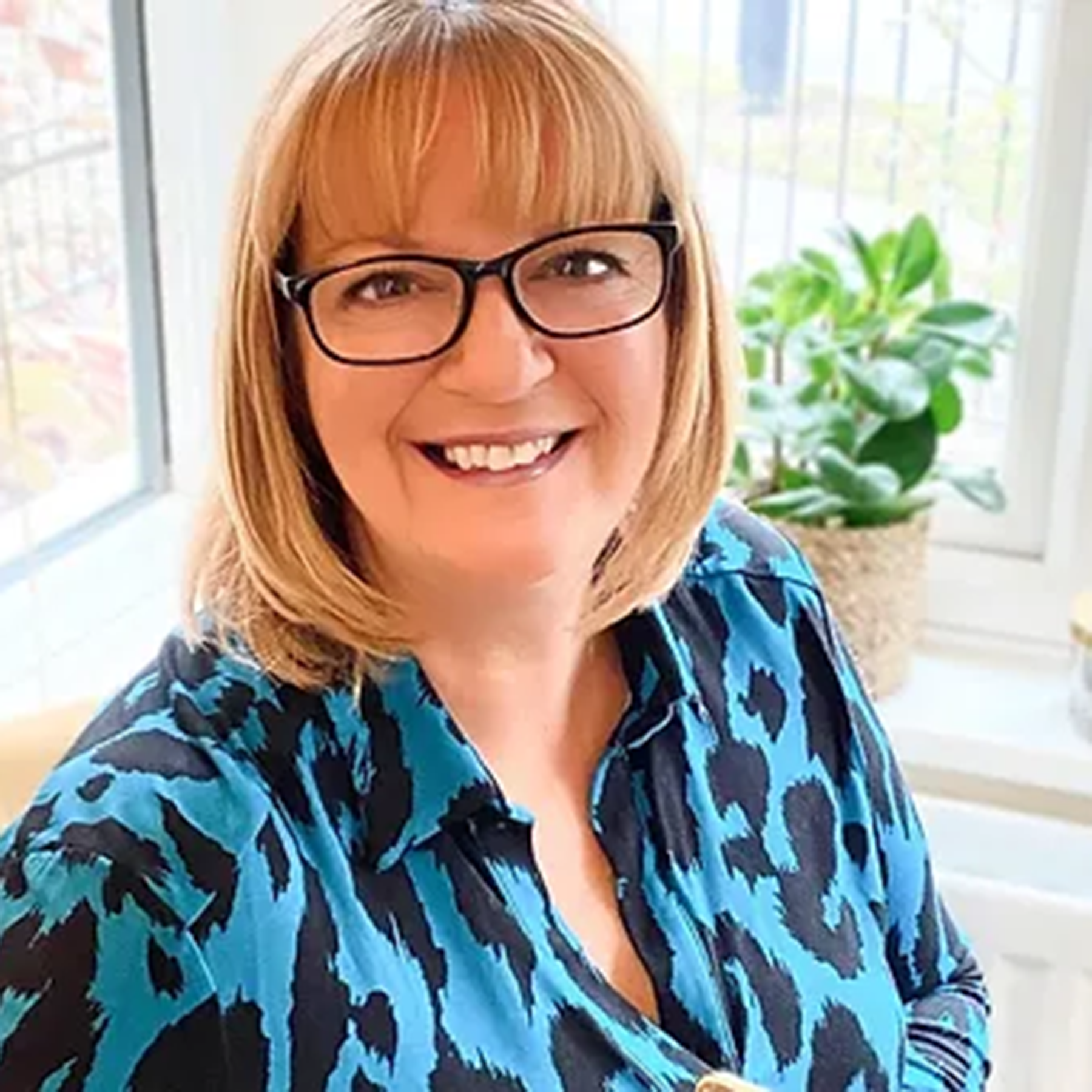 A woman wearing glasses and a blue shirt is smiling in front of a window.