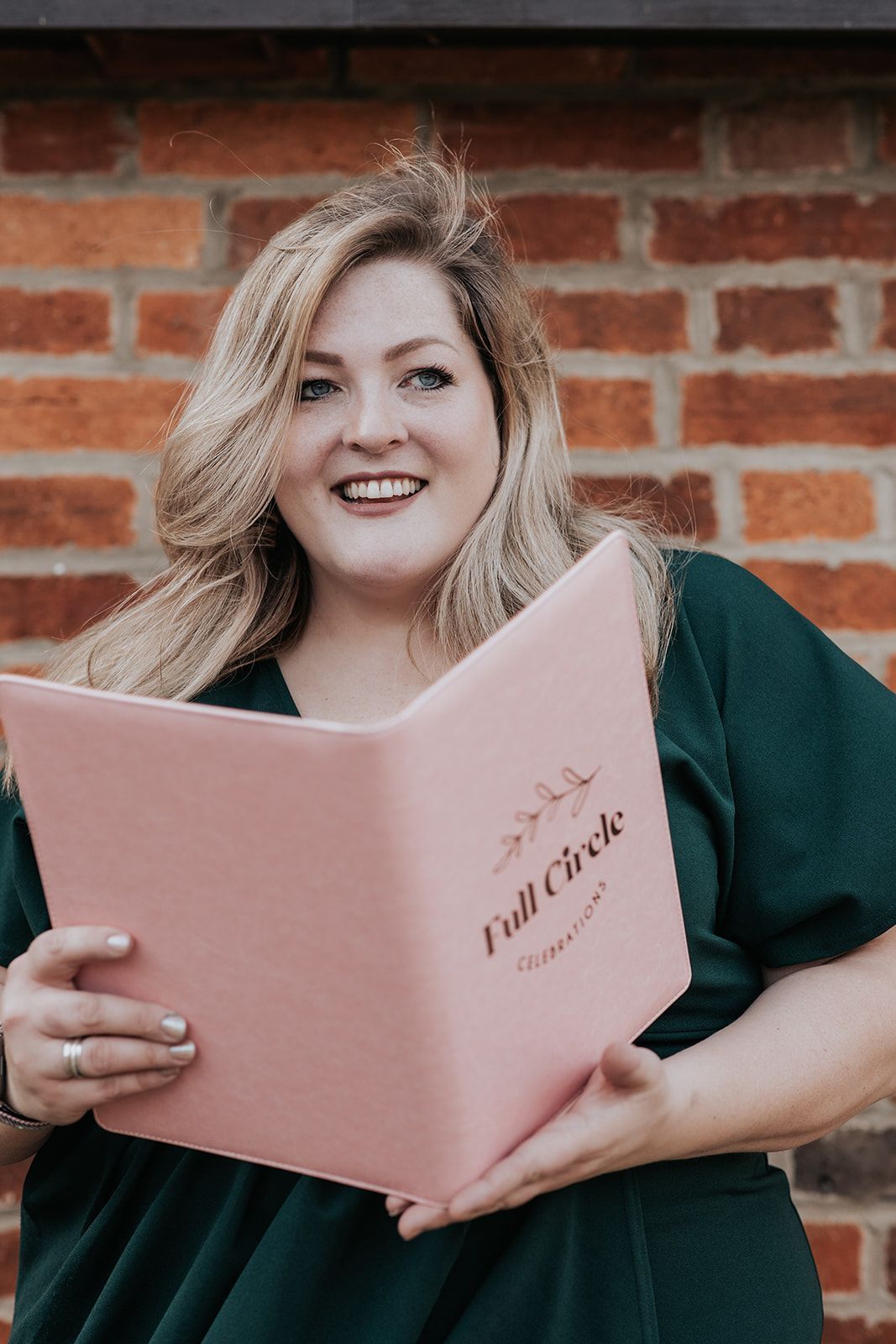 A woman in a green dress is holding a pink book in front of a brick wall.