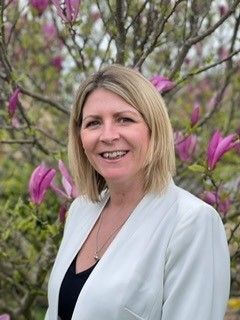 A woman in a white jacket is standing in front of a tree with purple flowers.