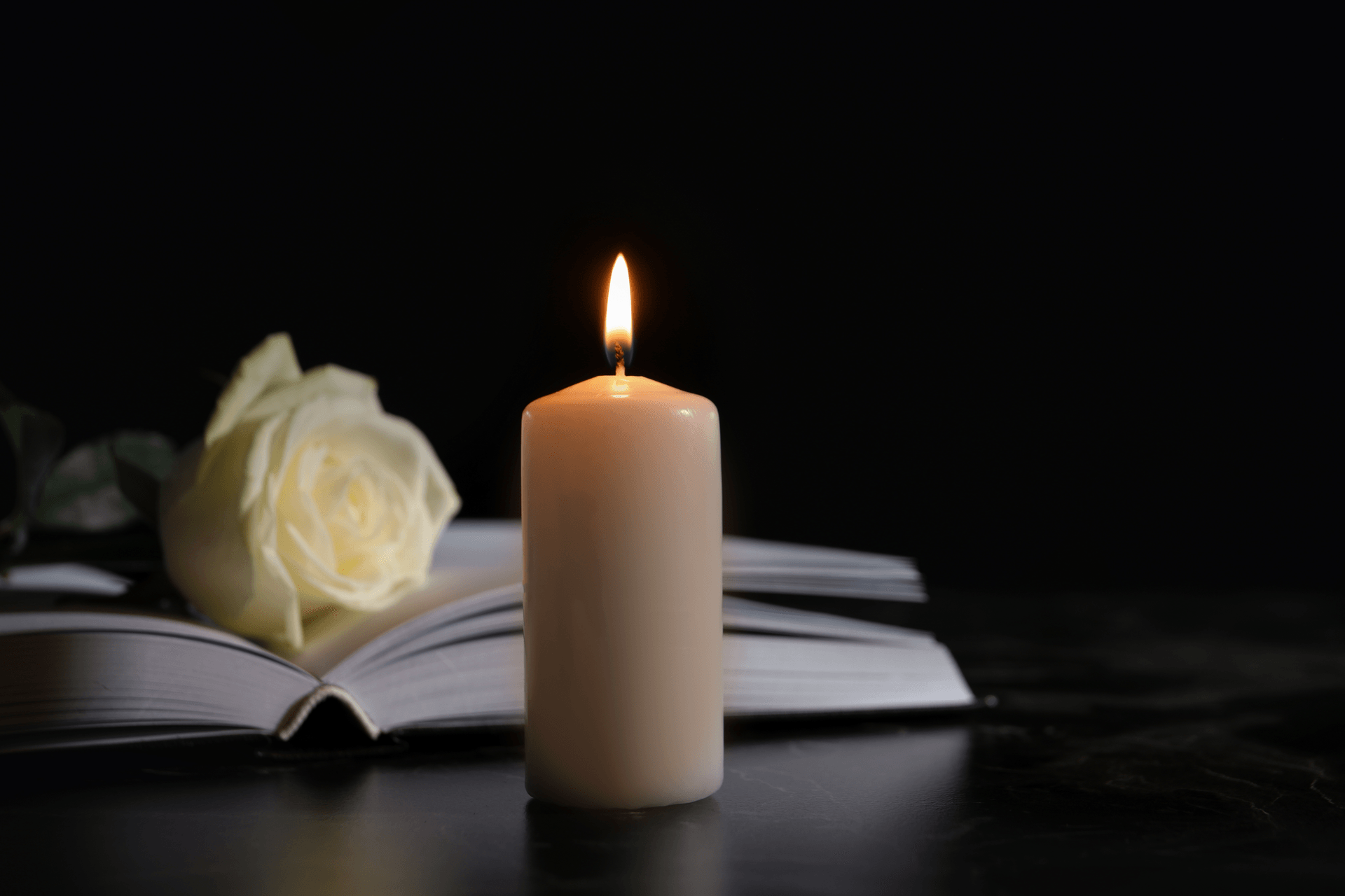 A candle is lit next to an open book and a white rose