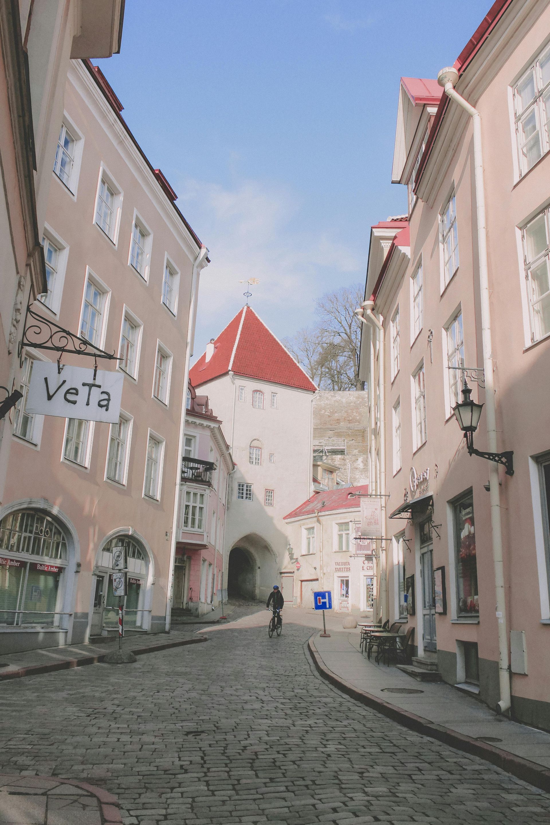 Cobblestone street lined with pink buildings, leading to a tower with a red roof, a cyclist rides.