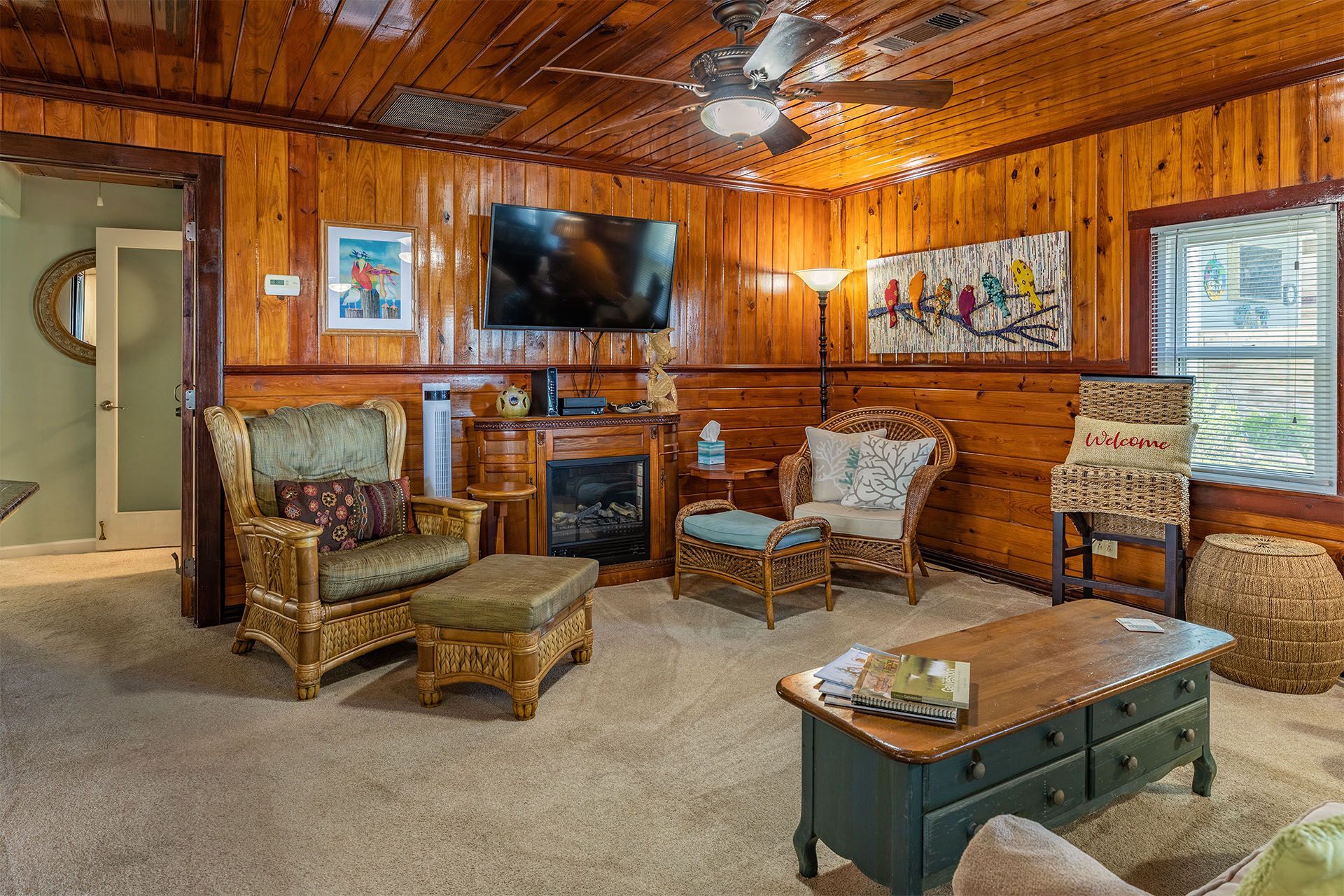 Cozy living room with wood paneling, rattan furniture, fireplace, TV, and artwork.