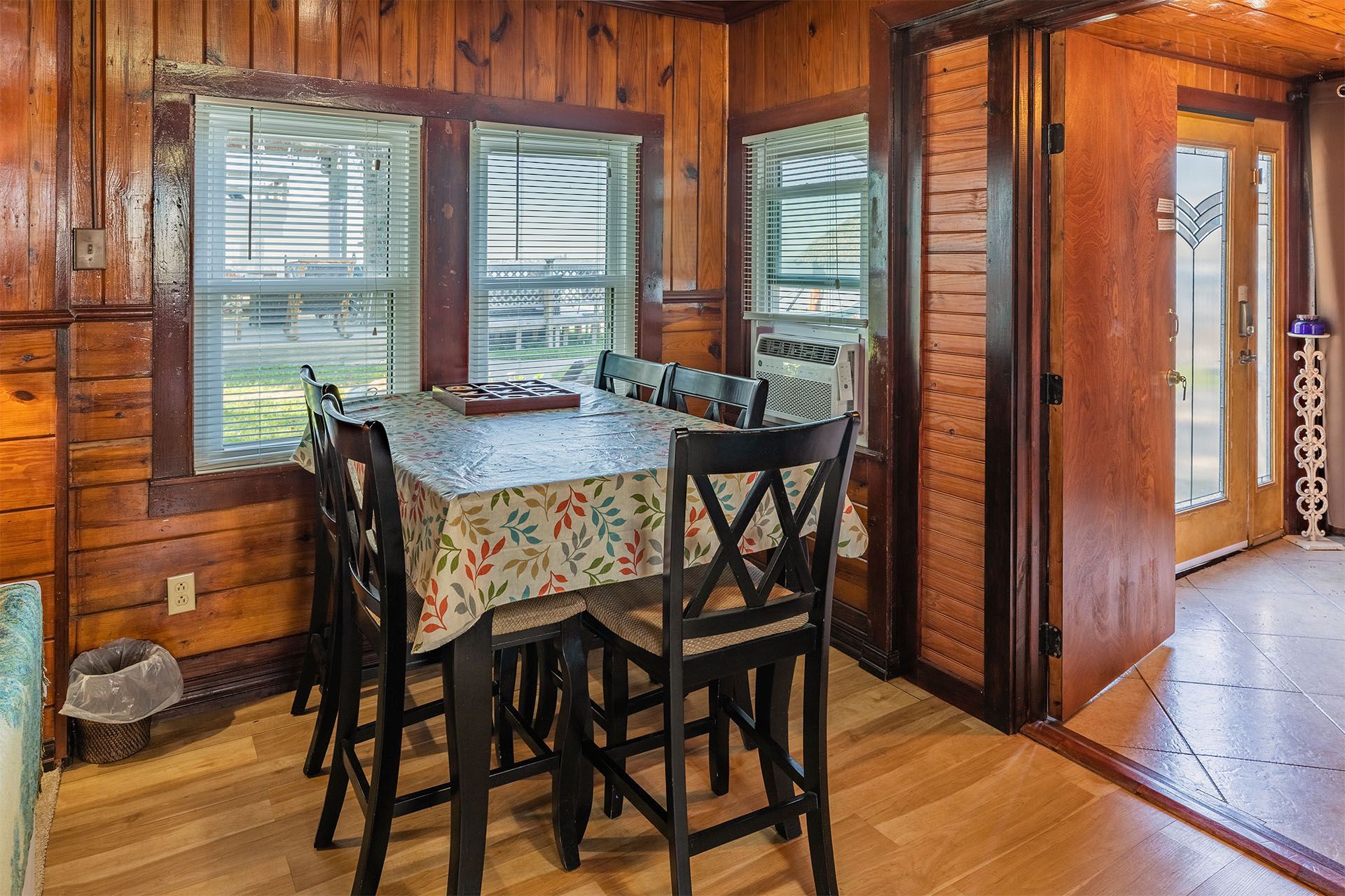 Dining area with a table set for six, wood paneled walls, and a view through the windows.