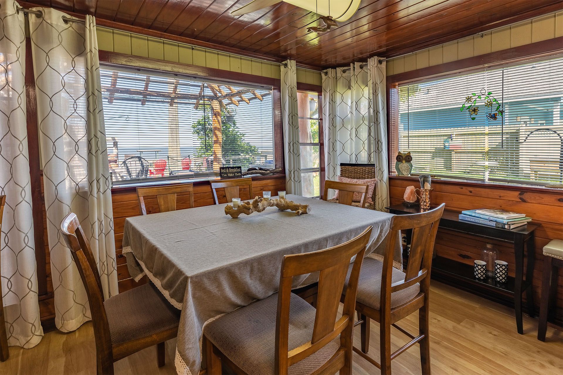 Sunroom with a wooden table set for dining, surrounded by chairs and windows with sheer curtains.
