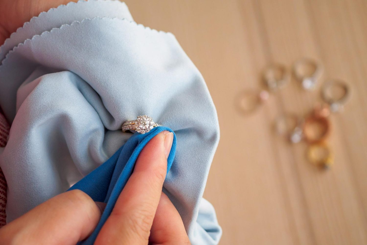 A person uses a light blue cloth to polish a diamond ring, with several other rings scattered on a wooden surface nearby.