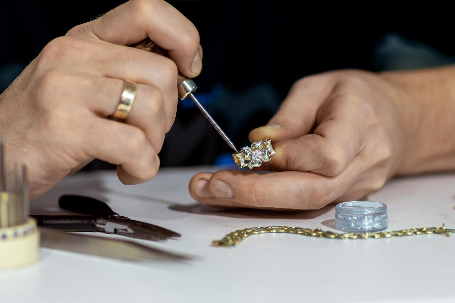 A jeweler works on a diamond ring with a small hand tool at a workbench with jewelry-making equipment.