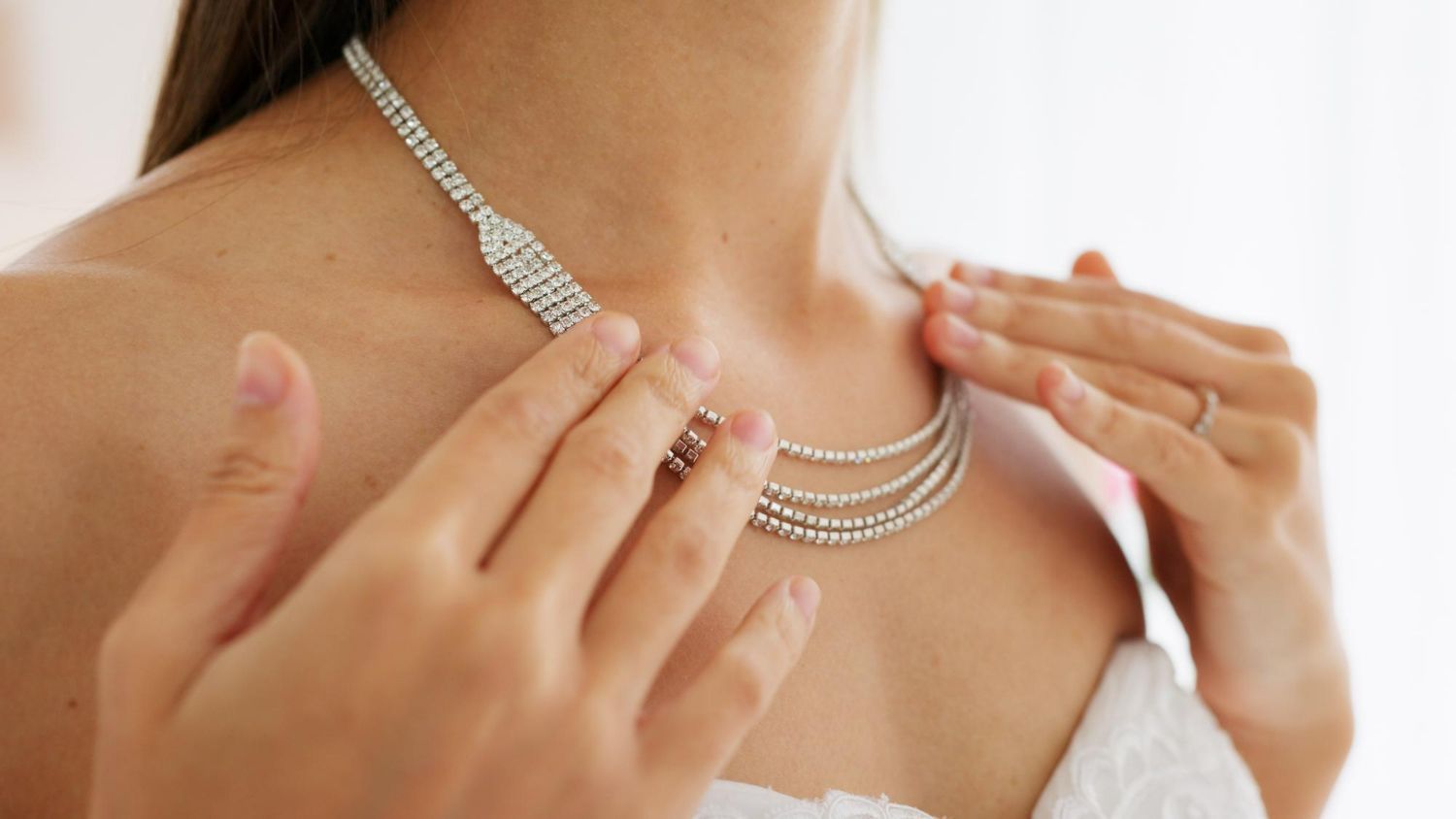 A person wears a multi-strand rhinestone necklace while adjusting it with their hands against their bare shoulders.