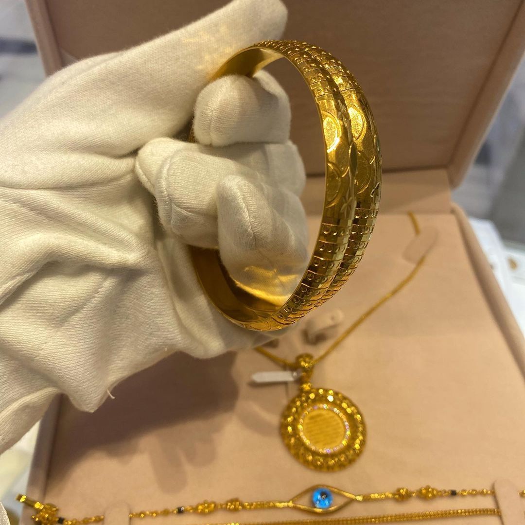 Gold bracelet held by gloved hand, with pendant necklace and bracelet in a jewelry box.