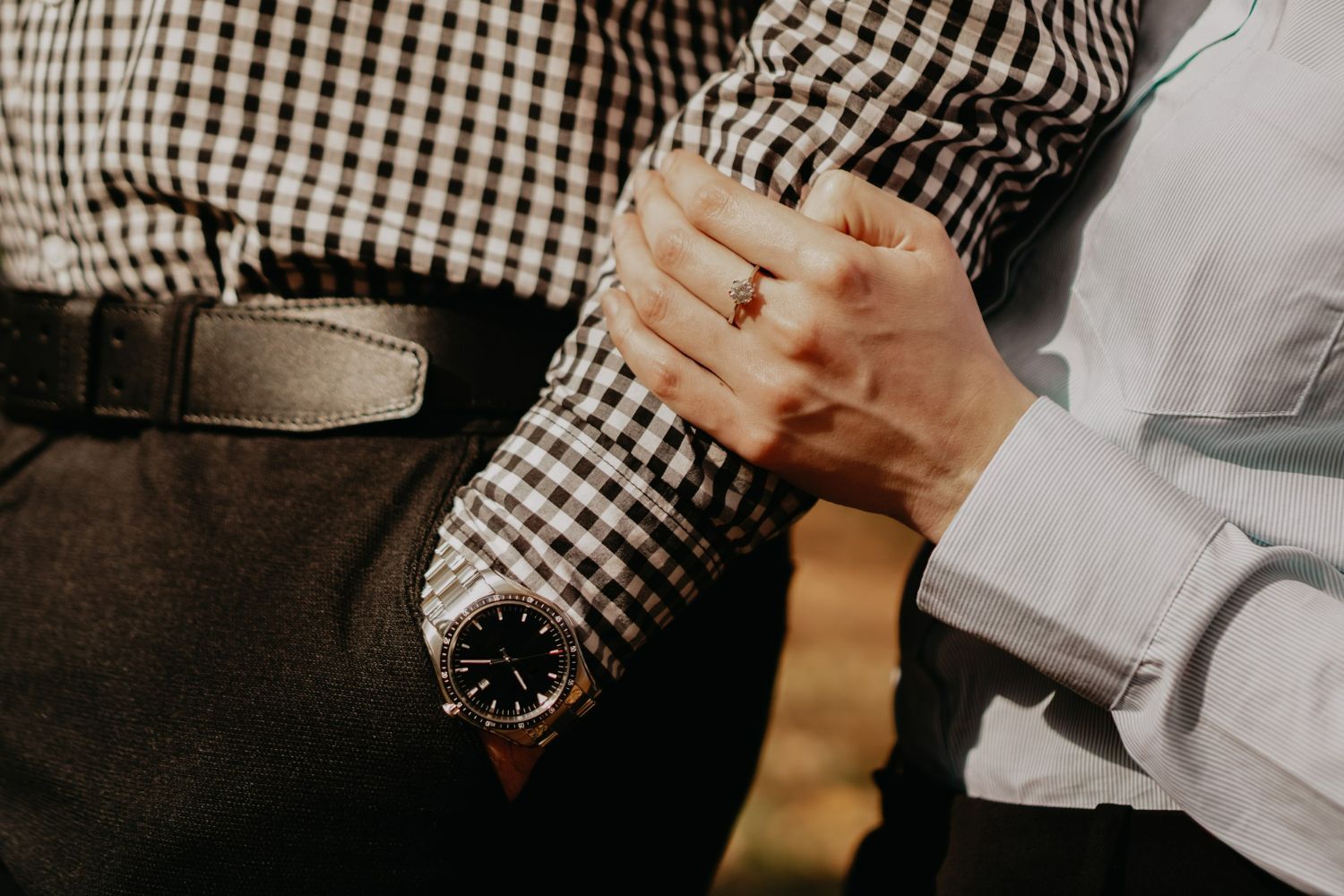 A person wearing a checkered shirt and watch with their hand gently held by another, showcasing a ring on their finger.