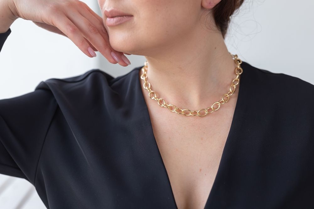 A person in a black V-neck top wears a thick gold chain-link necklace, resting their hand against their chin.