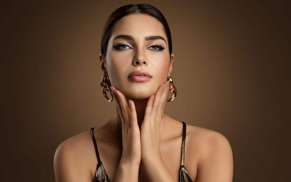 A person with dark hair and bold eye makeup poses with hands gently touching their face against a solid brown background.