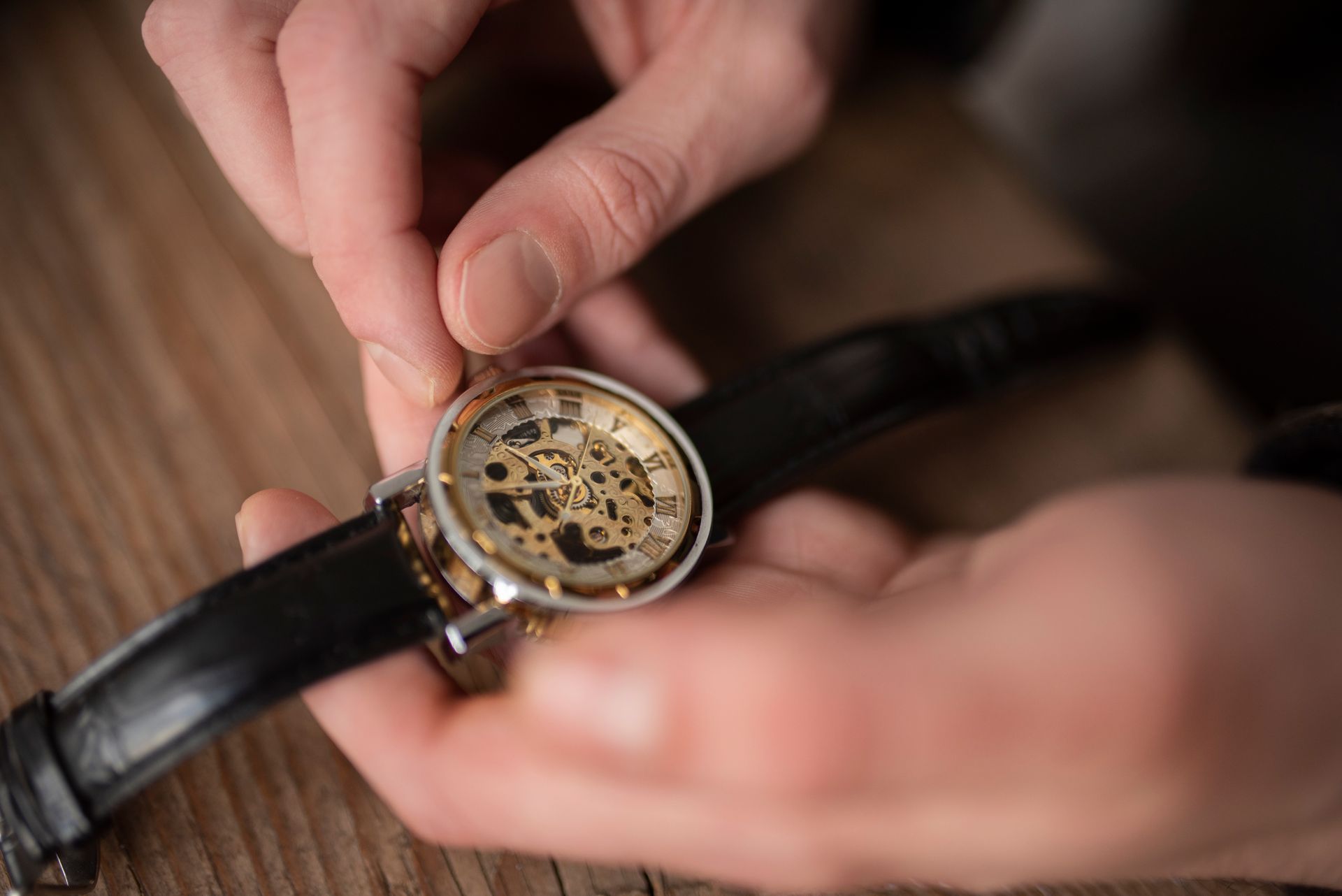 Hands adjusting the crown of a skeleton wristwatch with a black leather strap on a wooden surface.