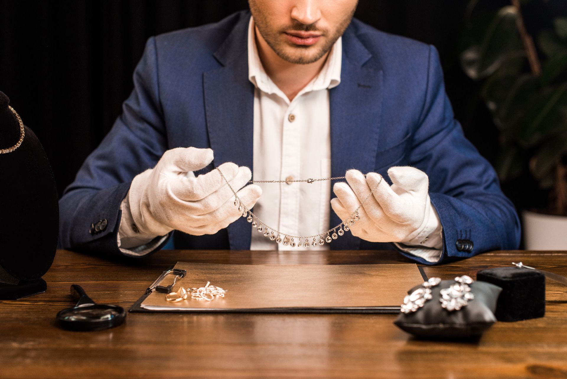 Jeweler in blue suit examines a diamond necklace with white gloves at a wooden desk.