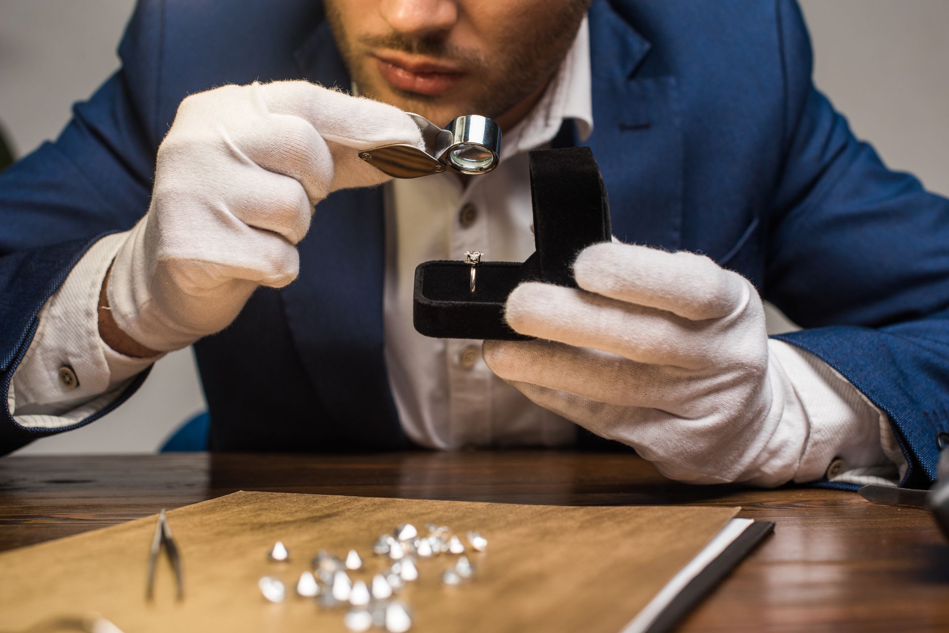 Jeweler examining a diamond ring with a loupe, wearing white gloves, with loose diamonds on the table.
