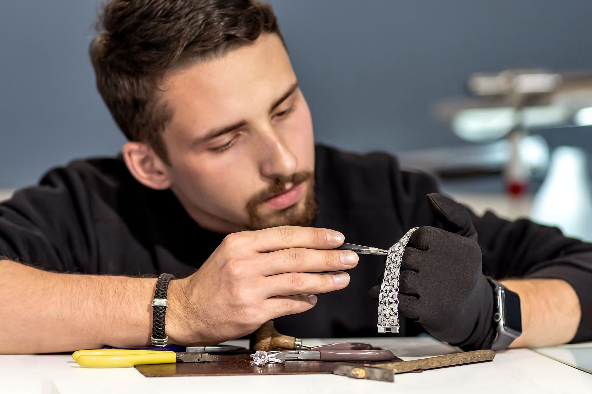 Man examining a silver watch bracelet with tools on a white surface, wearing a black glove.