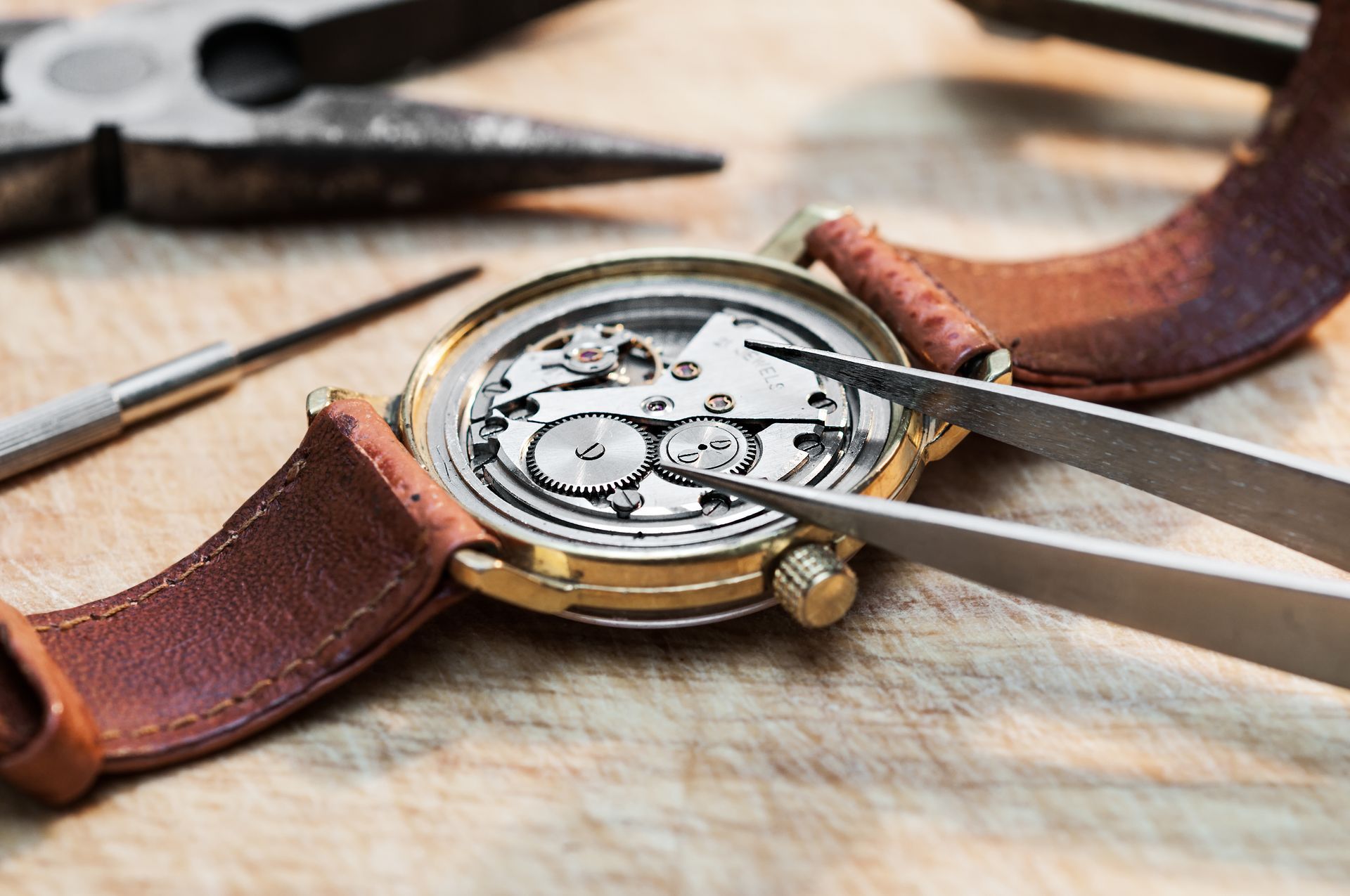 A watch is being repaired with tweezers on a wooden table.