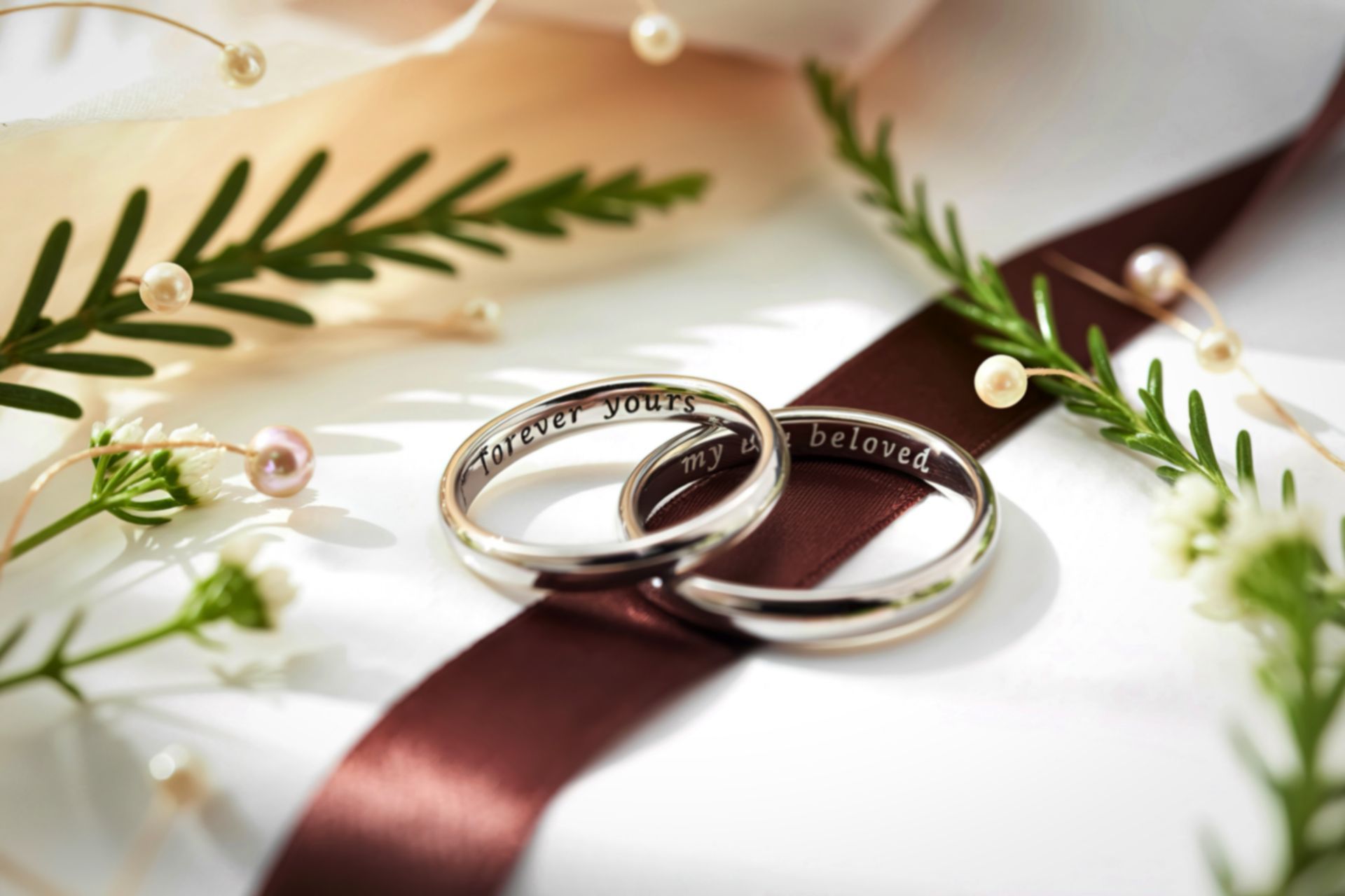 Two silver wedding rings on a dark brown ribbon, decorated with white flowers and pearls.