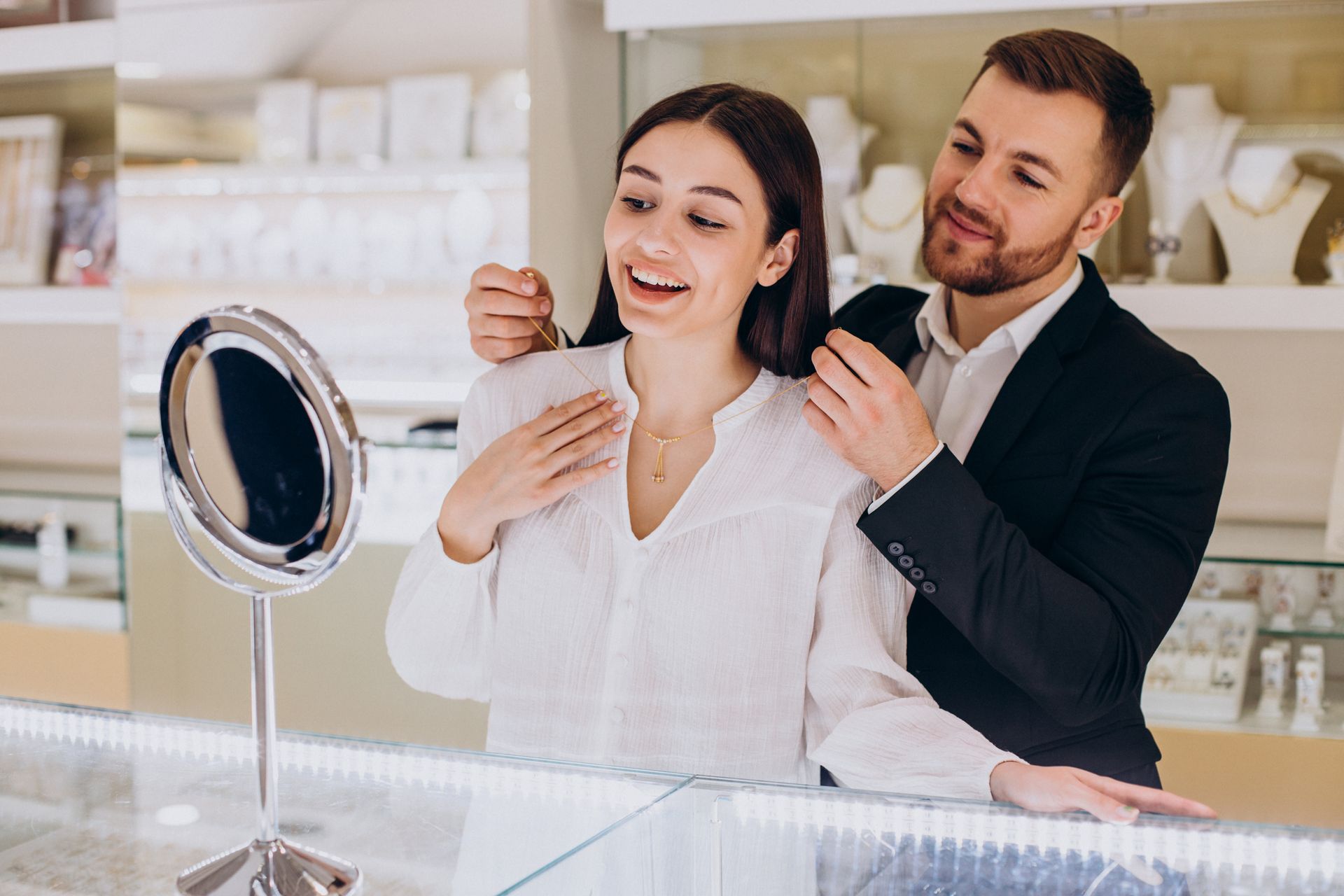 Man puts necklace on woman in jewelry store. They smile, looking in mirror at necklace.
