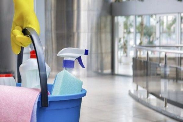 In un edificio moderno, una mano guantata di giallo regge un secchio blu pieno di flaconi di spray detergente e un panno rosa.