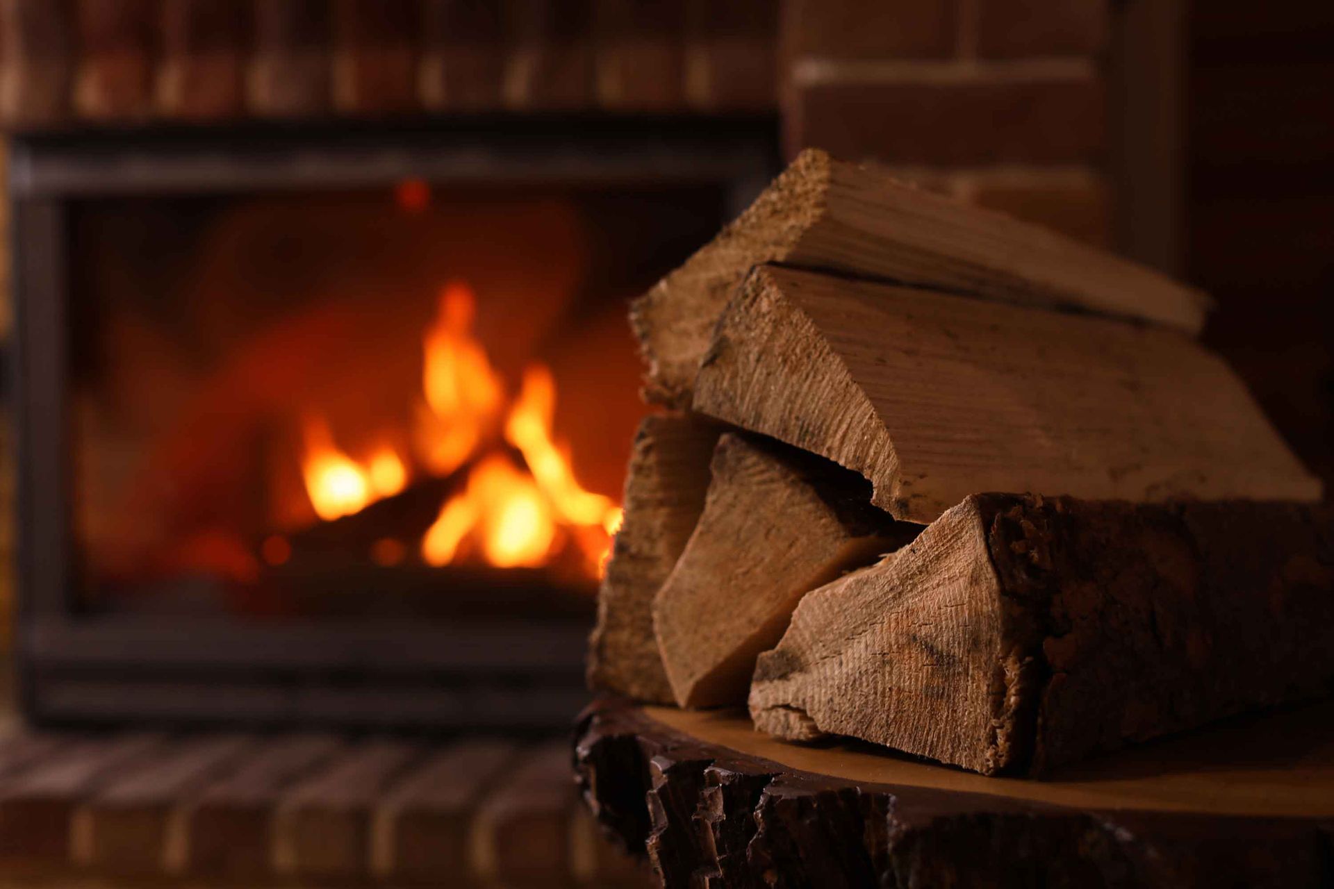 Close-up of a pile of wood, with a blurry and lit fireplace in the background.