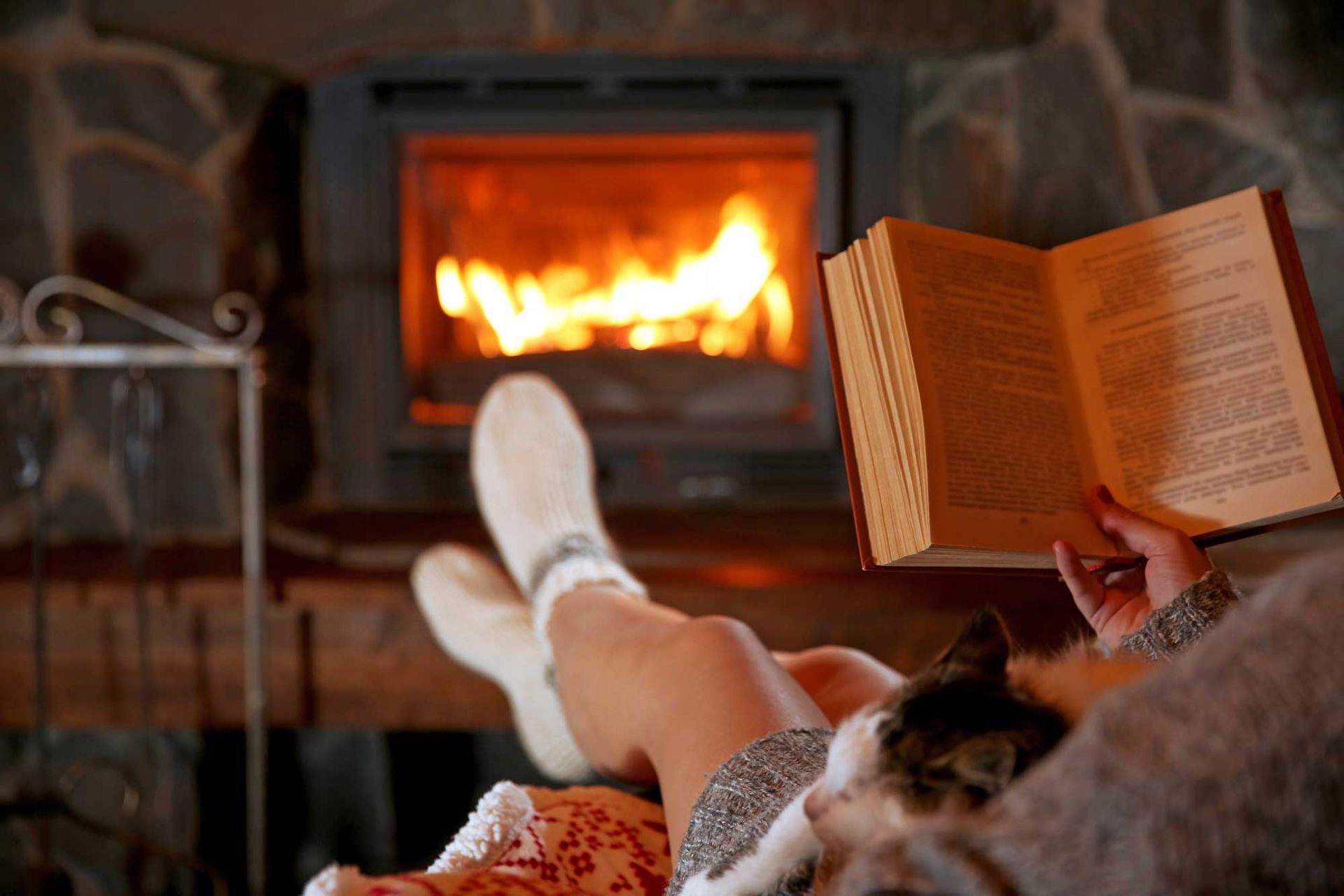 Person reading a book by a fireplace, cat on lap, feet in socks. Fire burning. Warm interior.