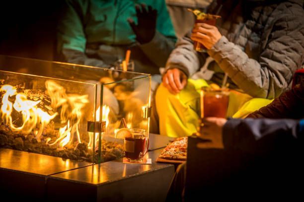 People gathered near a fire pit, holding drinks. Flames illuminate a clear glass barrier and the table.