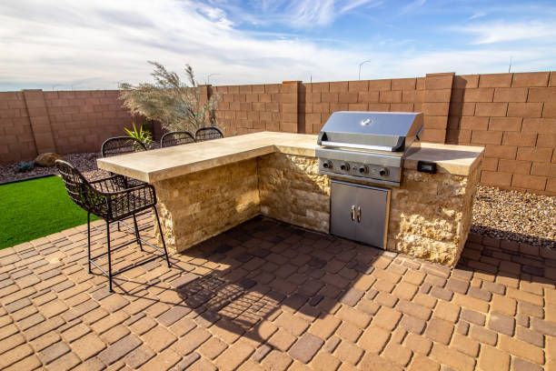 Outdoor kitchen with a built-in grill, stone counter, brick patio, and two bar stools.