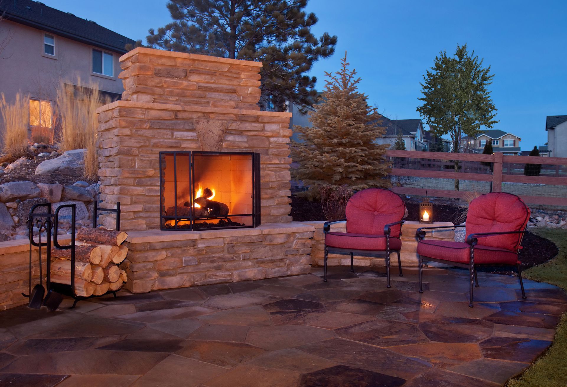 Stone fireplace with a burning fire, two red chairs, and a paved patio at dusk.