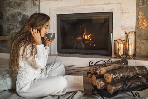 Woman in white holding a mug, sitting by a lit fireplace, reading. Firewood and candles nearby.