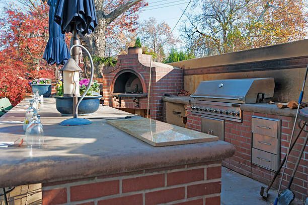 Outdoor kitchen with brick oven, grill, and countertop. Blue umbrella and fall foliage in the background.